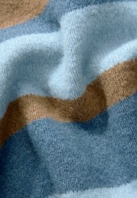 Tissu tricoté doux avec des rayures alternées en bleu clair, bleu foncé et marron, possédant une texture duveteuse et des détails de couture visibles.