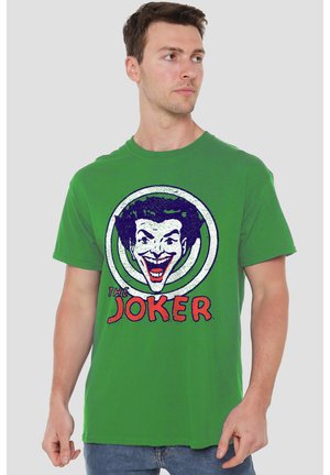 Mann trägt grünes T-Shirt mit großem Motiv eines lachenden Joker-Gesichts und dem Wort "Joker" in roten Buchstaben.