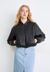 Μαύρο σακάκι από τεχνητό δέρμα με φερμουάρ μπροστά, κουκούλα και ελαστικές μανσέτες, συνδυασμένο με μια ανοιχτό μπλε τζιν φούστα με μπροστινή σχισ�μή.