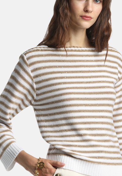 Femme portant un pull à rayures blanches et beiges à manches longues et un bracelet chaîne doré avec un charme rond au poignet gauche.