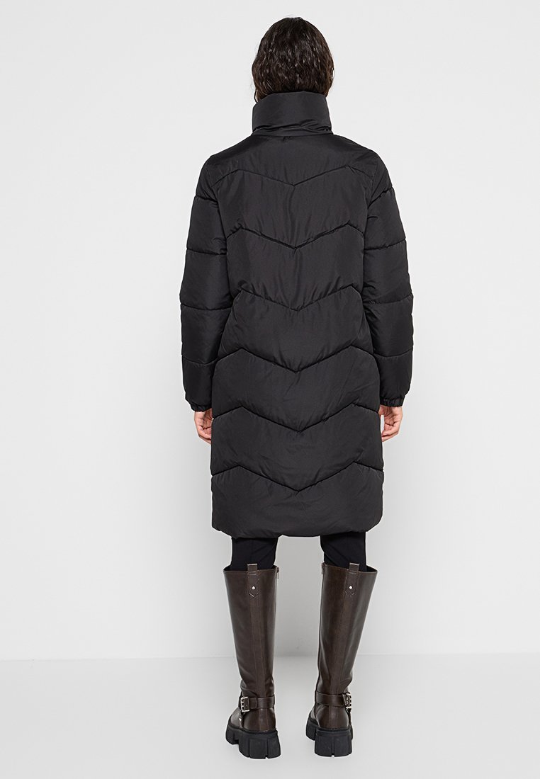 Langer schwarzer Puffer-Mantel mit hohem Kragen und Chevron-Steppnähten. Kombiniert mit knielangen braunen Stiefeln. Glatte Textur.