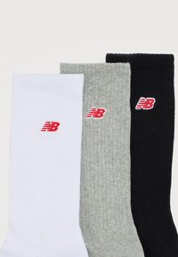 Τρεις καλτσες μερικώς ριγωτές σε λευκό, γκρι και μαύρο, καθεμία με ένα μικρό κόκκινο και λευκό λογότυπο «NB» κοντά στην κορυφή.