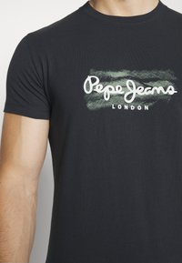 Camiseta de algodón negra con un logo blanco "Pepe Jeans" y "LONDON" sobre un fondo de pinceladas verdes. Diseño de manga corta y cuello redondo.