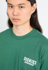 Zeleni pamučni majica s okruglim izrezom. Ima bijeli natpis "DICKIES WORKWEAR" na lijevoj strani prsnog dijela. Glatka tekstura, ležerni kroj.