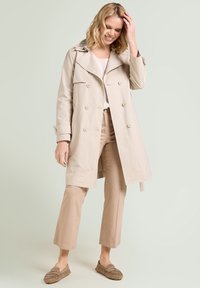 Mujer sonriente con cabello rubio, vistiendo un abrigo beige, pantalones claros, camisa blanca y mocasines marrones, de pie contra un fondo claro.