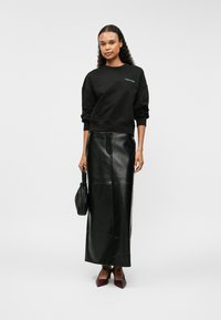 Μαύρη μπλούζα με στρογγυλή λαιμόκοψη και λογότυπο, συνδυασμένη με μακριά μαύρη δερμάτινη φούστα. Το μοντέλο κρατά μια μικρή μαύρη τσάντα και φορά μπορντώ τακούνια.