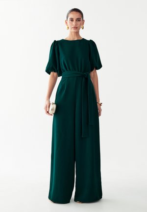 Femme portant une combinaison pantalon vert foncé à jambes larges avec manches bouffantes, taille nouée, tenant une petite pochette dorée et des boucles d'oreilles dorées sur un fond uni.