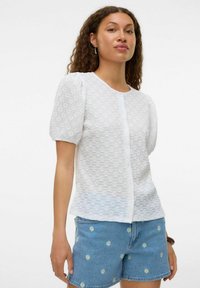 Blouse blanche à manches courtes avec un motif floral texturé, col rond et fermeture à boutons, associée à un short en denim bleu orné de broderies florales.
