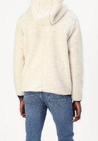 Vit fleecejacka med huva, texturerad yta och något sänkta axlar, som kombineras med blå jeans.