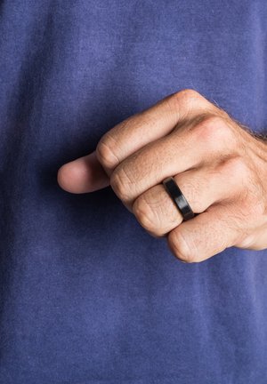 Black metal ring with a matte finish, featuring a flat profile, worn on the index finger of a hand against a purple fabric background.