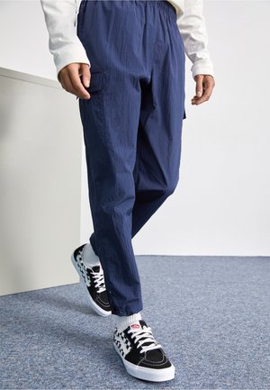 Personne portant un pantalon cargo bleu marine et des baskets à carreaux noirs et blancs marchant sur un tapis gris.