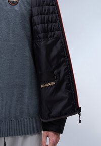 Személy kék kötött pulóvert visel, nyitott fekete puffert kabáttal, amelyen látható a belső címke és a piros cipzár szegély.