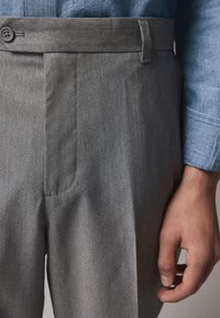 Pantaloni grigi su misura con una texture liscia, caratterizzati da una parte anteriore piatta e chiusura con bottone singolo, abbinati a una camicia celeste con bottoni.