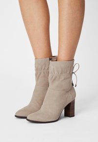 Bottes de cheville en suède beige avec un design décontracté, un bout pointu et un talon en bois de 3 pouces. Présente un accent à cordon au niveau du haut.