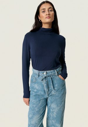 Femme aux longs cheveux foncés portant des boucles d'oreilles créoles dorées, un col roulé bleu marine et un jean taille haute bleu clair avec ceinture à nouer et poches.