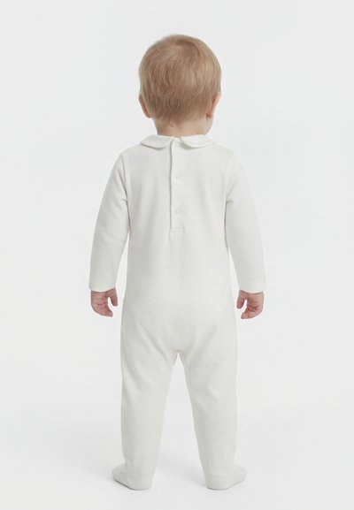 Småbarn står med ryggen mot, iført hvit heldrakt med lange ermer og føtter, med knapper bak og en Peter Pan-krage.