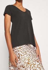 Frau trägt ein dunkelgraues, kurzärmeliges V-Ausschnitt-Oberteil und einen weißen Rock mit orangenen und braunen Punkten.