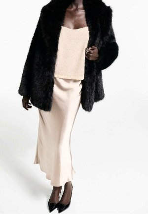 Personne à la peau foncée portant un manteau en fausse fourrure noire, un haut beige texturé, une jupe en satin beige et des chaussures à talons aiguilles noires en marchant.