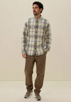 Beige geruite flanellen shirt met blauwe accenten, twee borstzakken en een paarse broek, gecombineerd met grijze sneakers. Zachte textuur en knoopsluiting.