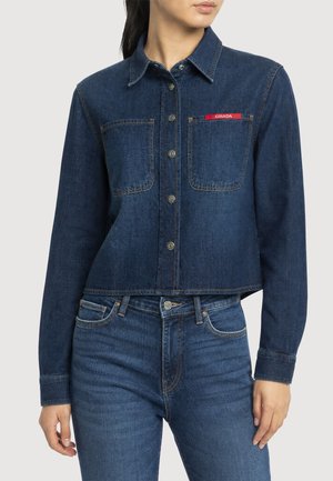 Femme portant une veste en denim bleu foncé avec poches avant et boutons, assortie à un jean bleu foncé sur fond uni.