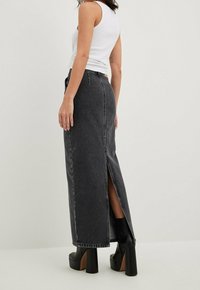 Mujer vestida con una falda larga de mezclilla negra de cintura alta con abertura en la parte trasera, blusa sin mangas blanca y botas de plataforma negras, de pie de lado.