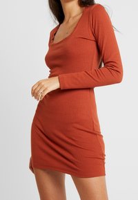 Robe à manches longues côtelée en orange brûlé, présentant un décolleté arrondi et une silhouette ajustée. Le matériau est doux avec un léger stretch.