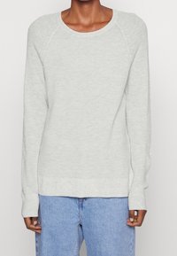 Helles grau gestricktes Sweatshirt mit einem runden Ausschnitt, langen Raglanärmeln und gerippten Manschetten. Kombiniert mit hellblauen Jeans.