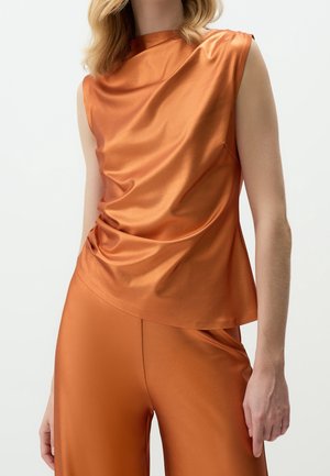 Femme portant un haut sans manches en satin orange brillant et un pantalon large assorti sur fond blanc uni.