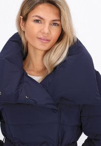 Młoda kobieta z blond włosami, ubrana w ciemnoniebieską kurtkę puchową z dużym kołnierzem, na jednolitym białym tle.