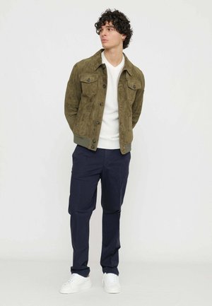 Giacca in suede oliva con chiusura a bottoni, due tasche sul petto e orlo a costine; abbinata a pantaloni blu navy e sneaker bianche. Sfondo neutro.