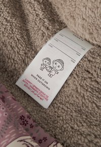 Myk beige fleece-stoff med en vaskeanvisningsetikett som inneholder illustrasjon, tekst og et fargerikt blomster mønster på den nedre kanten.