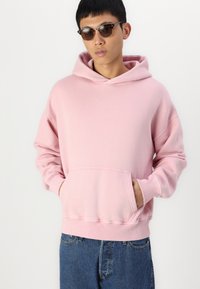 Sweatshirt à capuche rose clair avec une poche avant, coupe ample et poignets côtelés. Associé à un jean en denim bleu foncé et des lunettes de soleil.