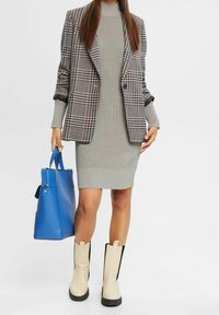 Blazer a scacchi con accenti blu navy, vestito grigio a coste, stivali beige fino al ginocchio con lati neri e borsa a tracolla blu.