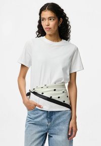 Vit kortärmad t-shirt tillsammans med en krämfärgad midjescarf med prickar, prydd med ett svart band och svarta detaljer. Ljust blå denimjeans.