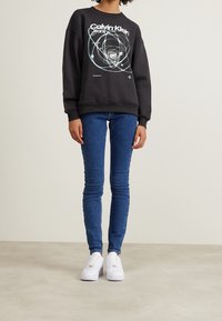 Svart bomullströja med grafiskt tryck och ribbade muddar. I kombination med blå tighta jeans och vita sneakers. En enkel bakgrund.