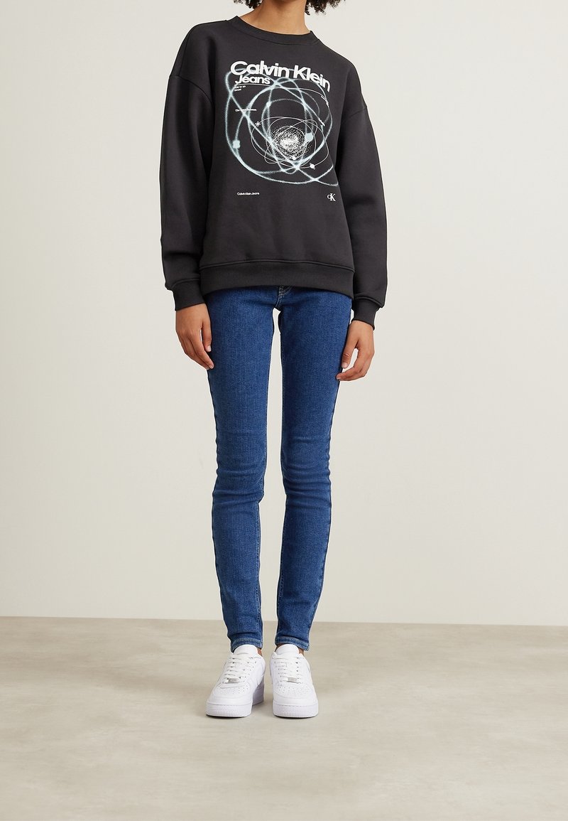 Svart bomullströja med grafiskt tryck och ribbade muddar. I kombination med blå tighta jeans och vita sneakers. En enkel bakgrund.