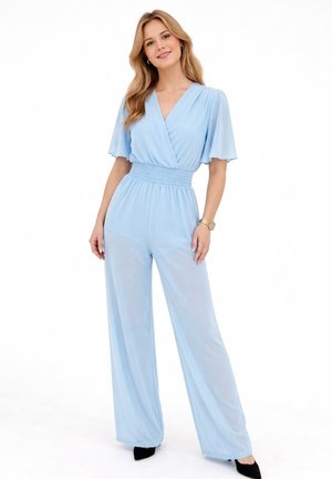 Kobieta o długich blond włosach, ubrana w jasnoniebieski kombinezon szerokimi nogawkami z falbanowymi rękawami, w czarnych, szpiczastych butach, stojąca i uśmiechająca się.