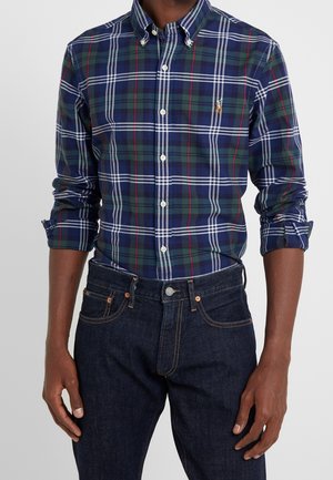 Mand iført langærmet ternet skjorte i blå, grøn og hvid med ærmerne rullet op og mørkeblå jeans.