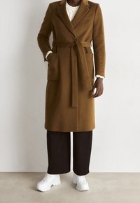 Cappotto marrone in misto lana con cintura annodata, colletto a rever e tasche frontali, abbinato a pantaloni neri ampi e sneakers bianche.