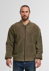 Oliwkowa polarowa kurtka z zamkiem błyskawicznym z przodu, wysokim kołnierzem i bocznymi kieszeniami. Luźny krój z miękką teksturą i ściągaczami przy mankietach.