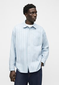 Άνδρας που φοράει ανοιχτό μπλε πουκάμισο με κάθετες ρίγες και σκουρόχρωμο παντελόνι, κοιτάζοντας προς τα δεξιά του με ένα χέρι στην τσέπη.