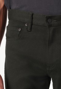 Jeans verde oscuro de mezclilla, con cierre de botón y cremallera, bolsillos delanteros y detalles de costura en contraste.