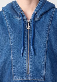 Sweat à capuche en denim bleu avec fermeture éclair à l'avant et cordons bleus, montrant la partie supérieure de la poitrine et du cou d'une personne qui le porte.