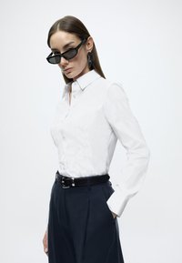 Λευκό πουκάμισο με κουμπιά και γιακά, συνδυασμένο με σκούρο παντελόνι και μαύρη ζώνη. Αξεσουάρ περιλαμβάνουν μαύρα γυαλιά ηλίου και εντυπωσιακά σκουλαρίκια.