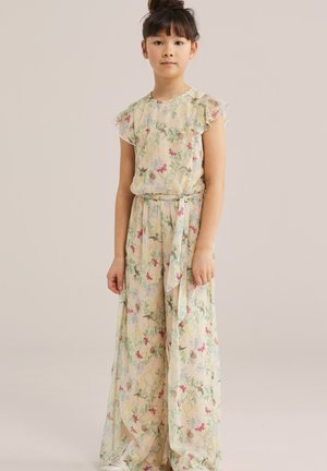 Ragazza giovane con capelli scuri raccolti in uno chignon, indossa un abito lungo giallo chiaro con motivi floreali e farfalle, in piedi davanti a uno sfondo beige.