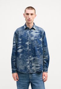 Blauwe spijkerblouse met een patroonontwerp, voorzien van een knoopsluiting aan de voorkant, kraag, borstzak en lange mouwen. Geweven stof.