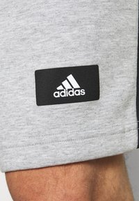 Pantalones cortos deportivos grises con una textura suave, con un parche del logo de Adidas negro cosido en el lateral. Los detalles incluyen un borde oscuro.