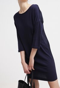 Abito blu navy a maniche corte realizzato in tessuto morbido, con una vestibilità sciolta, scollatura rotonda e polsini arrotolati, abbinato a una borsa nera.