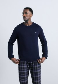 Άνδρας που φοράει μπλε σκούρο πουκάμισο με μακριά μανίκια και μαύρο-μπλε καρό παντελόνι, ρυθμίζει το τελείωμα του πουκαμίσου, κοιτάζοντας ελαφρώς αριστερά πάνω σε μονόχρωμο φόντο.