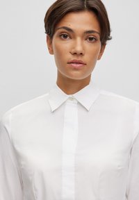 Camicia bianca con colletto classico e maniche lunghe, realizzata in tessuto liscio e leggero con una leggera lucentezza.